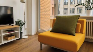 A modern living room with a yellow sofa, a flat-screen TV, a floor lamp, a potted plant, and a view of a brick building through a window. - MEL&BENS Domshof | City center | Balcony | (Bremen)