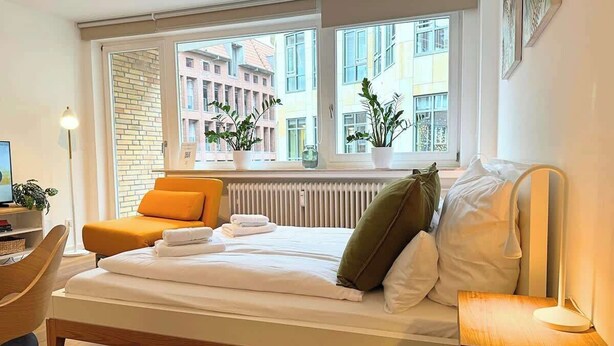 A hotel room with a bed, a chair, a desk, a lamp, and a window with a view of buildings. - MEL&BENS Domshof | City center | Balcony | (Bremen)