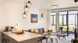 Cozinha moderna com ilha central, área de jantar com mesa e cadeiras, e sala de estar com sofá e poltronas. Uma grande janela oferece vista para o exterior.