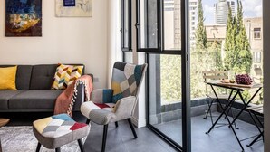 Uma sala de estar moderna com um sofá, duas poltronas e uma mesa com frutas. Janelas amplas oferecem vista para edifícios e árvores.