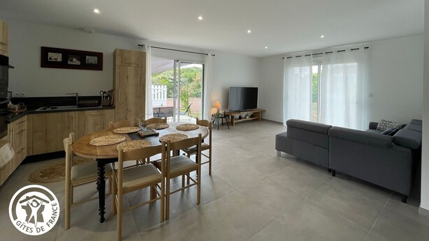 A modern living room with a grey sofa, a dining area with a wooden table and chairs, a kitchen with wooden cabinets, and a TV on a stand. - Charming 6-Person Cottage Near Ocean & Cycling Routes (Beauvoir-sur-Mer)