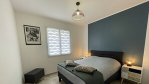 A bedroom with a bed, bedside tables, a nightstand, a picture frame, and a window with patterned blinds. - Charming 6-Person Cottage Near Ocean & Cycling Routes (Beauvoir-sur-Mer)