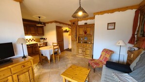 A cozy living room with a sofa, a coffee table, and a TV. There is a kitchen area with wooden cabinets and a dining table. - Cozy 2-BR Apt for 6 Near Pool, Ski Bus & Amenities (Les Carroz d'Arâches)