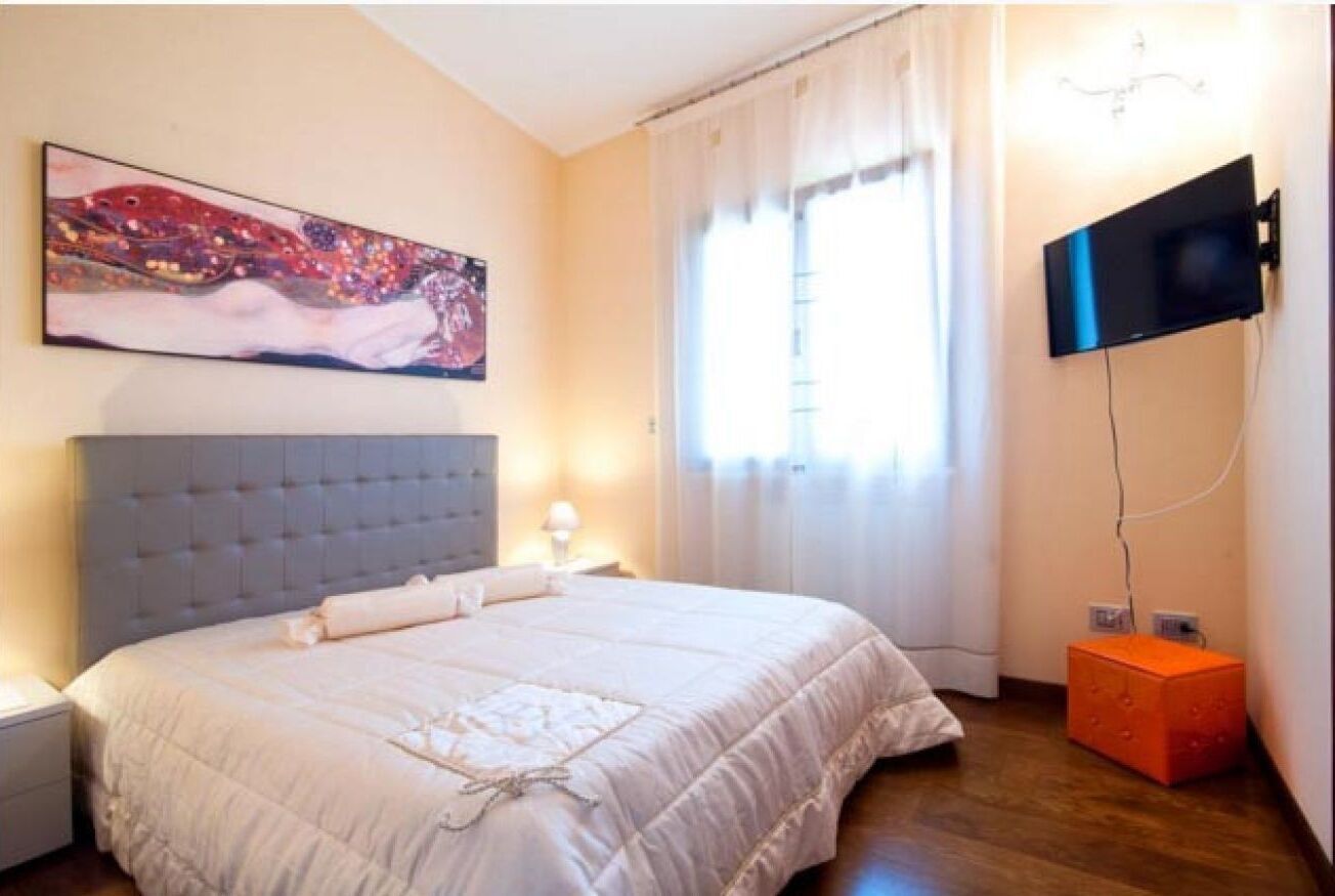 Ein Schlafzimmer mit einem großen Bett, einem flachbildfernseher an der Wand, einem Gemälde über dem Bett und einem Fenster mit Vorhängen.