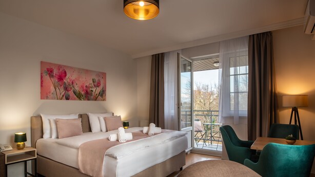 A bedroom with a large bed, a desk, and a chair. There is a window with curtains, a painting on the wall, and a lamp on the nightstand. - Holdvirág Hotel (Sarvar)