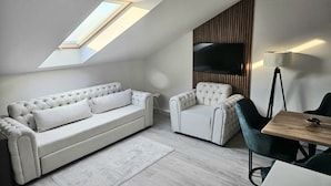 A modern living room with a high ceiling, a sofa, a chair, a small table, and a TV mounted on the wall. - Holdvirág Hotel (Sarvar)