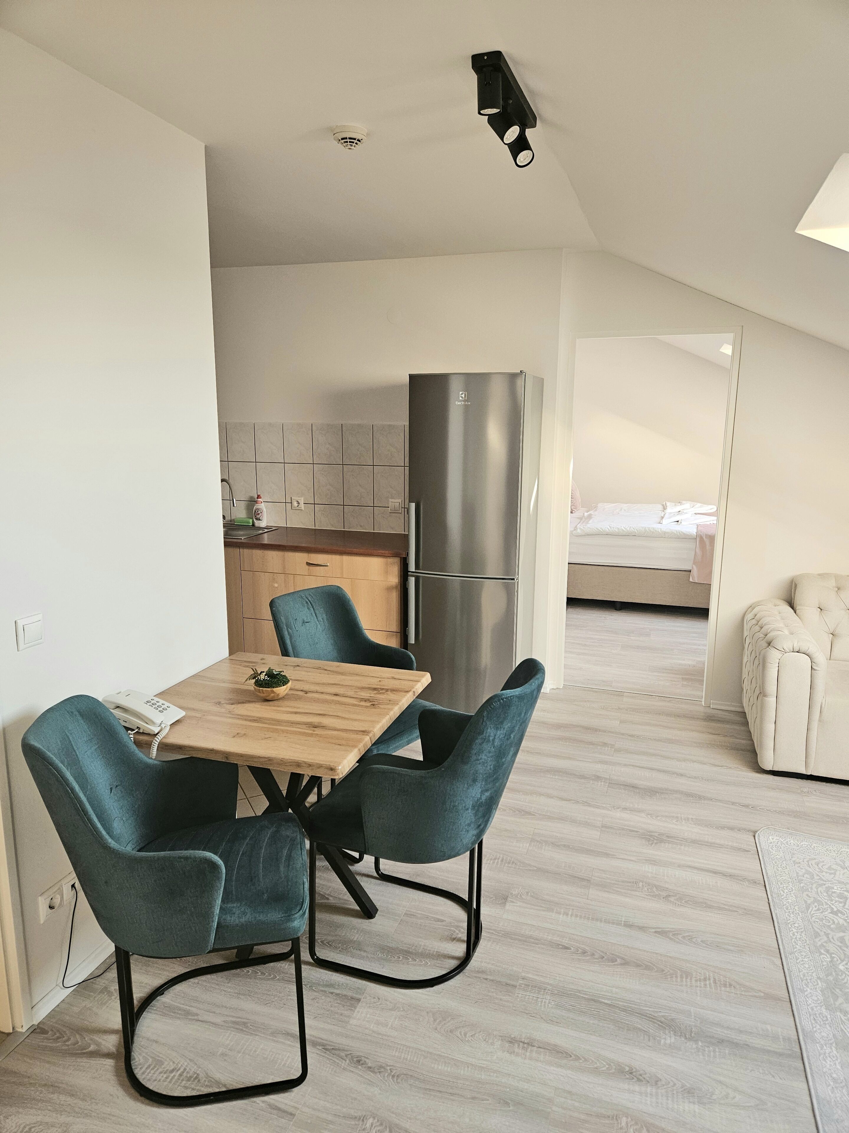 A modern kitchen with a stainless steel refrigerator, a dining area with a wooden table and teal chairs, and a bedroom visible through an open door.