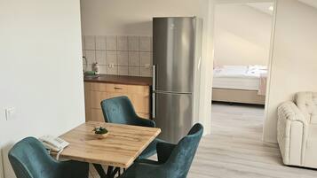 A modern kitchen with a stainless steel refrigerator, a dining area with a wooden table and teal chairs, and a bedroom visible through an open door.