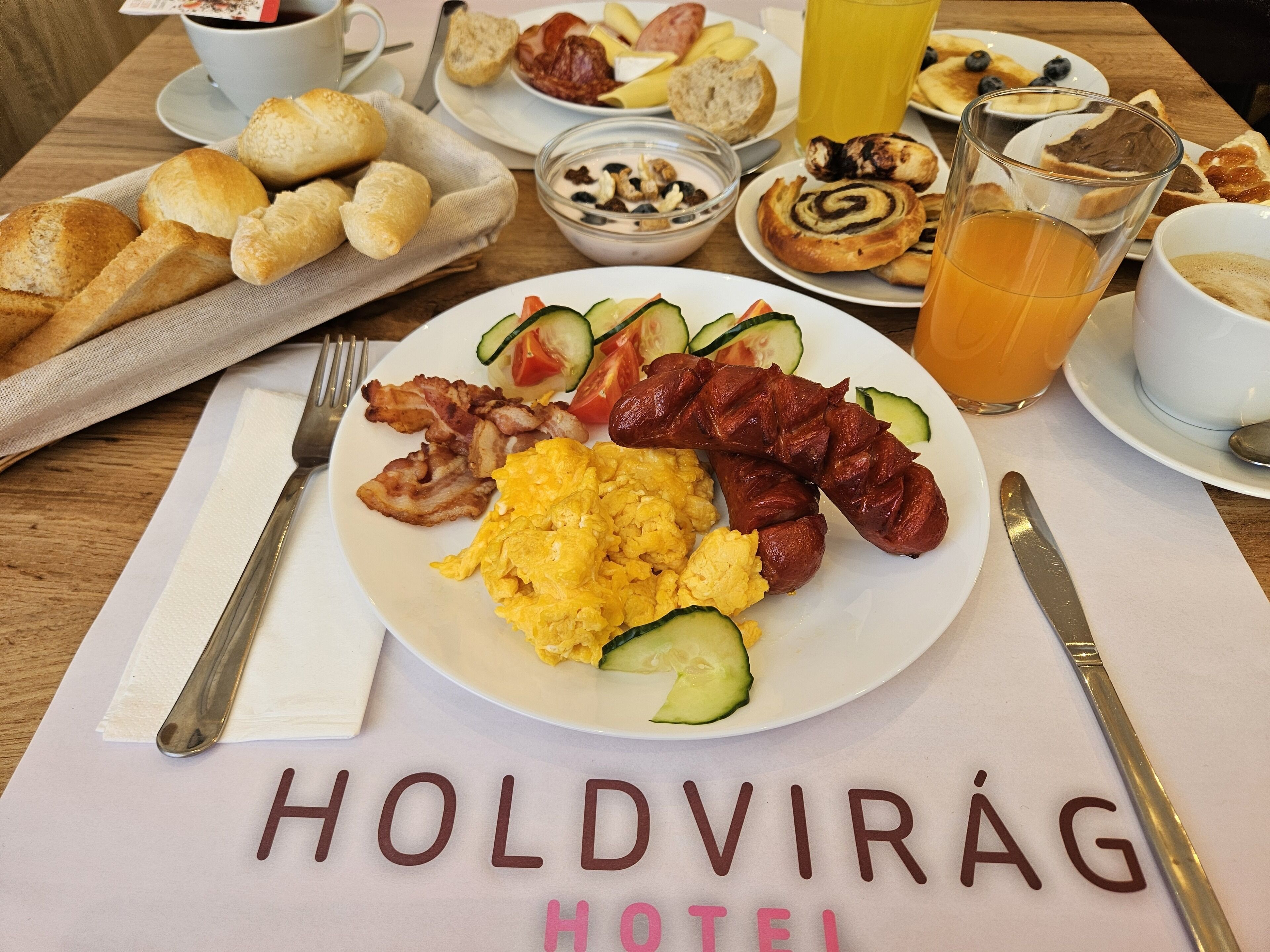 A breakfast plate with eggs, bacon, sausage, and vegetables, along with a glass of orange juice and a cup of coffee on a table with a placemat reading "Holdvirág Hotel".