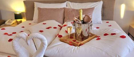 Quarto de hotel com uma cama decorada com pétalas de rosa, uma garrafa de champanhe e duas taças.