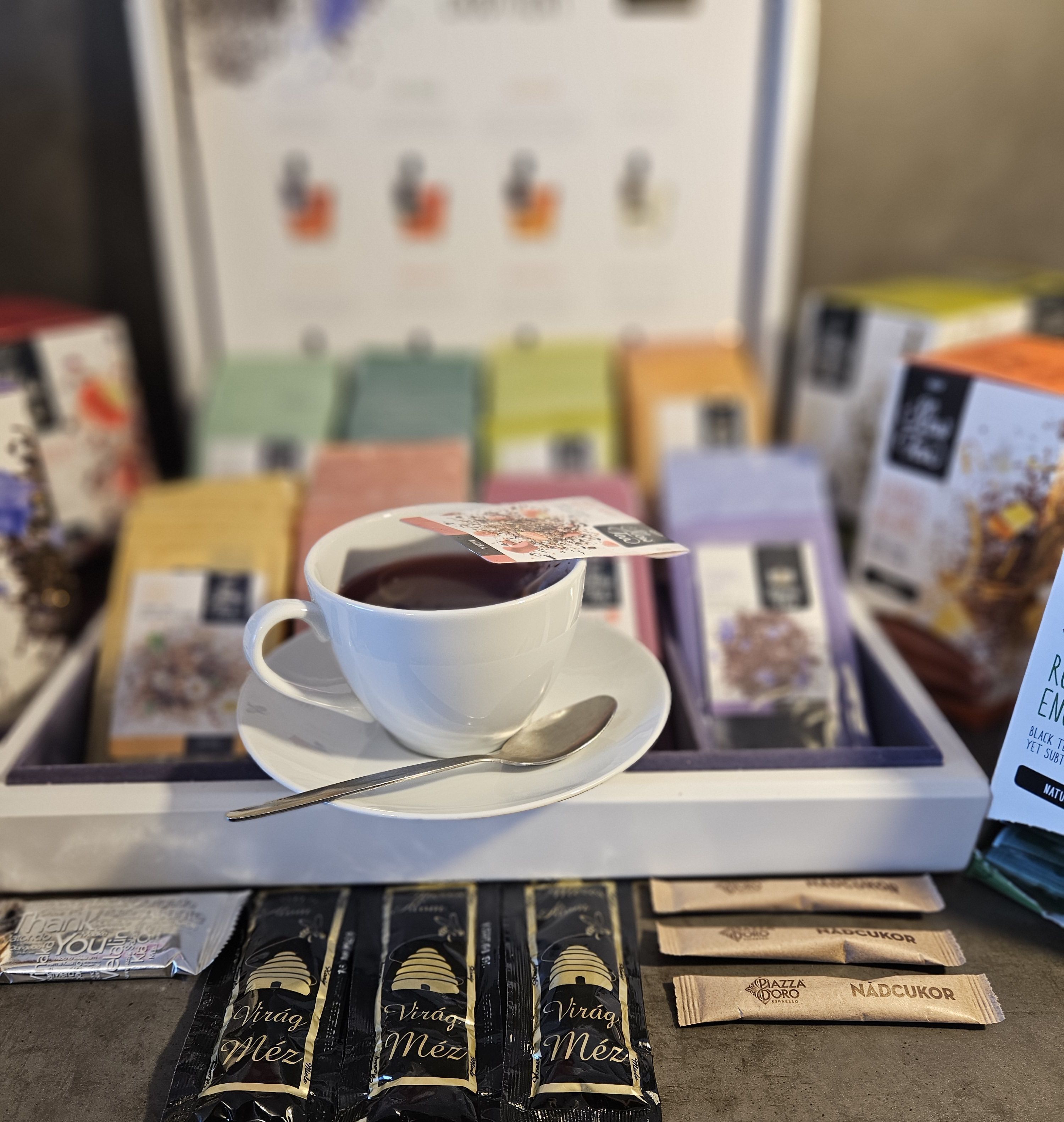 A cup of coffee on a tray with several coffee pods labeled 'Virág Méz' and 'NADCUKOR' in front of a colorful display of coffee pods.