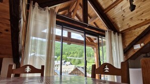 Ein Holzinnenraum mit Tisch und Stühlen, großen Fenstern mit Vorhängen und Blick auf einen bewaldeten Bereich.