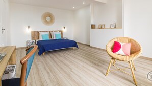 A spacious bedroom with a wooden floor, a bed with a blue bedspread, a wicker chair with cushions, and a wall-mounted mirror. - Family house near the beach (Saint-Marcouf)