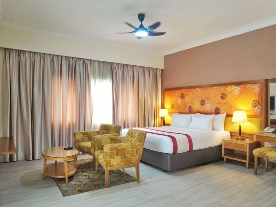 A hotel room with a bed, two bedside lamps, a ceiling fan, a sofa, a small table, and a chair.