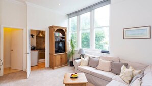 A living room with a sofa, a coffee table with fruit, a bookshelf, and a window with a view of greenery. - Charming Studio Apartment Near Clapham Common (London)