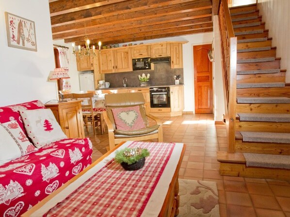 Ein gemütliches Wohnzimmer mit einem roten, gemusterten Sofa, einem Holzesstisch und einem Sessel mit einem herzförmigen Kissen. Im Hintergrund ist eine Küche mit Holdekor und einem Herd erkennbar.