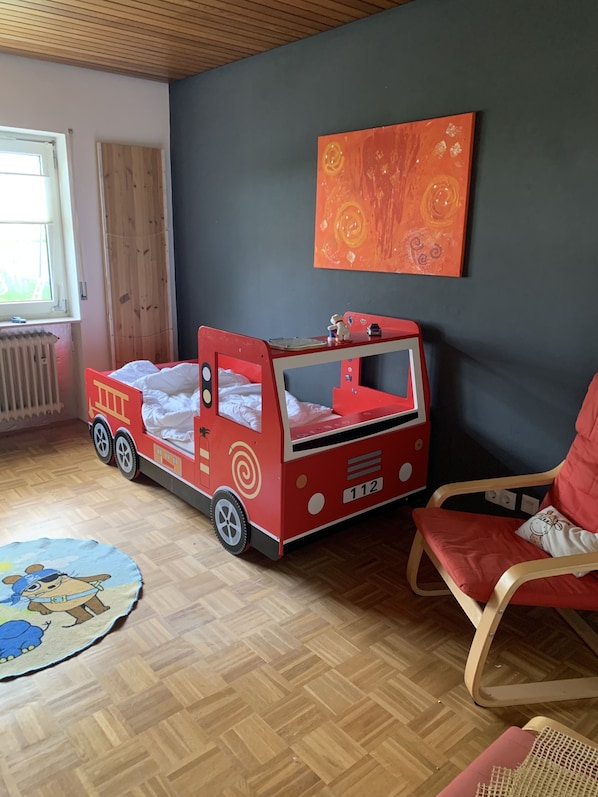 Une chambre d’enfant avec un lit rouge, un fauteuil à bascule, un radiateur et une fenêtre.