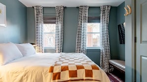 Ein Schlafzimmer mit einem großen Bett, einem karierten Bettbezug, punktierten Vorhängen, einer hölzernen Bank und einem gemusterten Teppich.