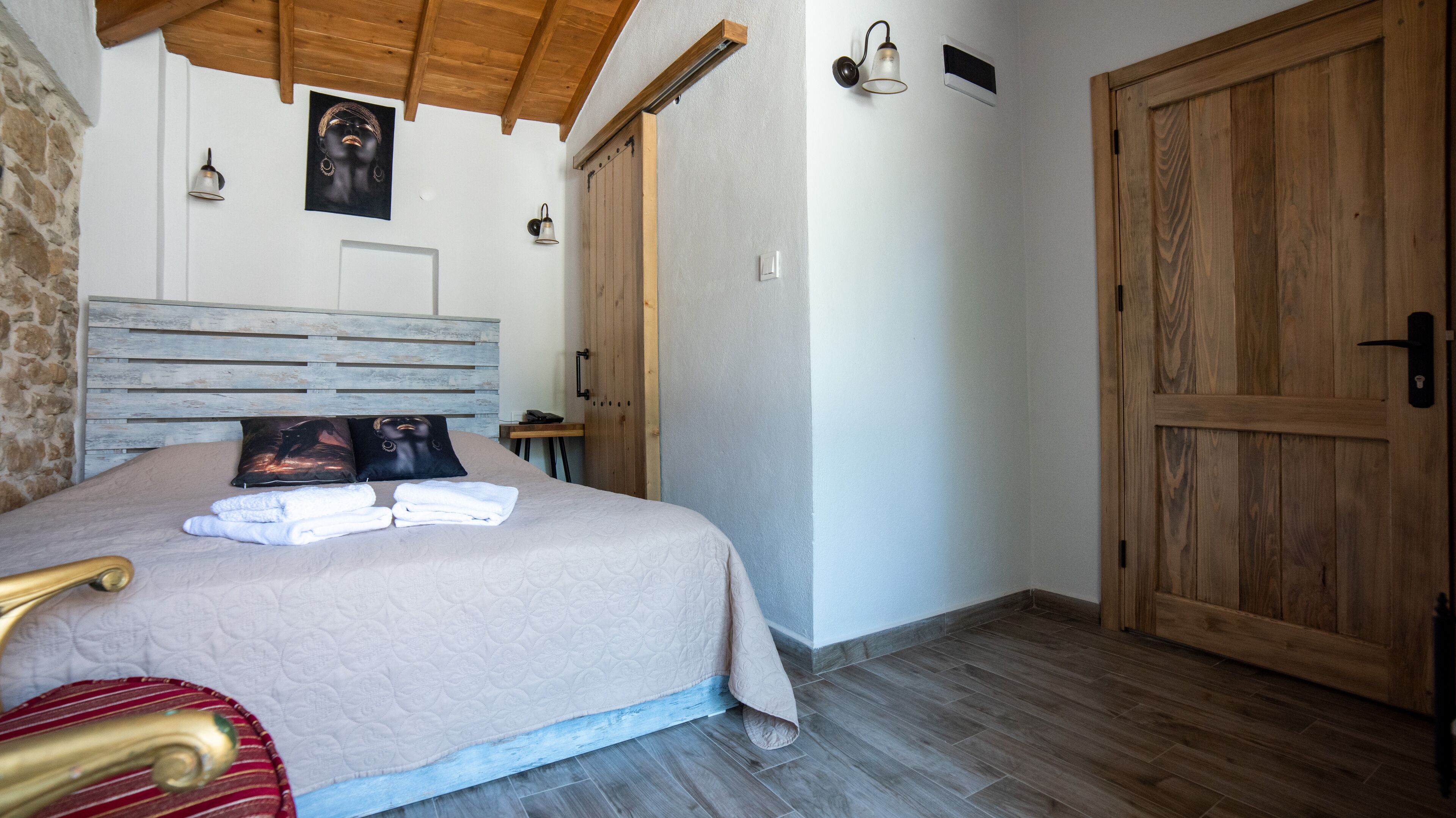 Ein Schlafzimmer mit einem hölzernen Kopfteil, einem Bett mit beiger Bettdecke, zwei Kissen, einem Nachttisch, einer Holztür und einer Steinwand.