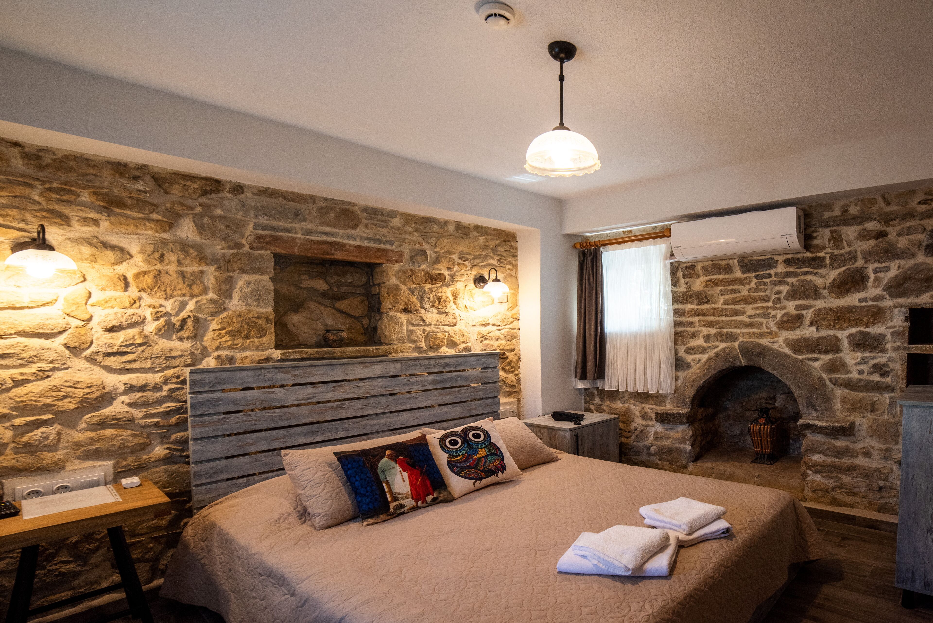 Ein Schlafzimmer mit Steinwand, hölzernem Kopfteil, einem Bett mit beiger Decke, zwei Kissen, einem Nachttisch, einer Lampe und einem Kamin.