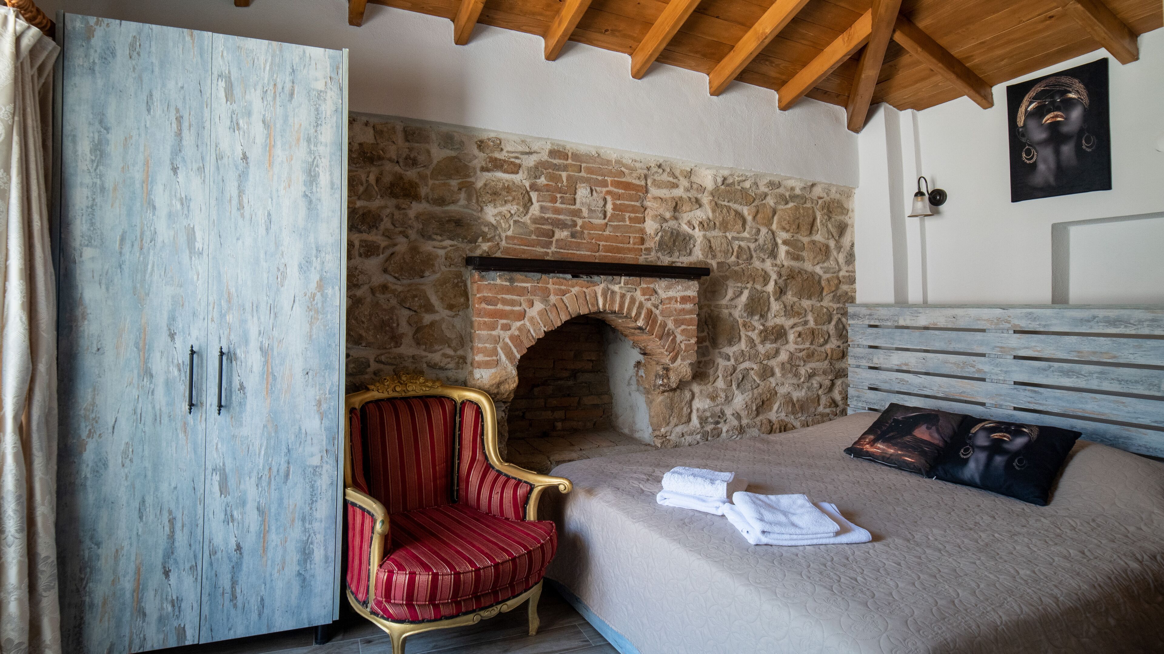 A bedroom with a stone fireplace, a red upholstered chair, a bed with a white comforter, and a wooden headboard.