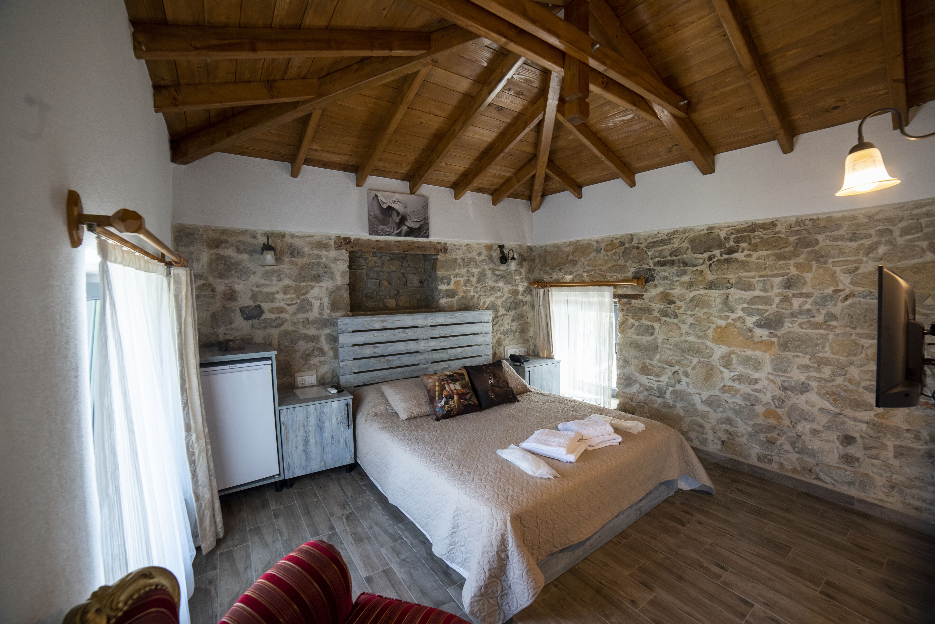 Quarto com parede de pedra, vigas de madeira, cama com manta bege, mesa de cabeceira pequena e cadeira vermelha.