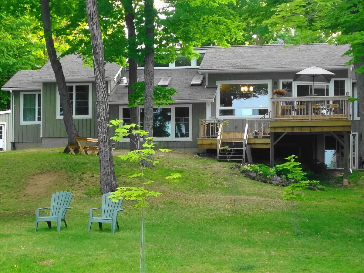 Une maison à deux étages avec une terrasse et un espace extérieur couvert. On y trouve deux chaises Adirondack dans le jardin.