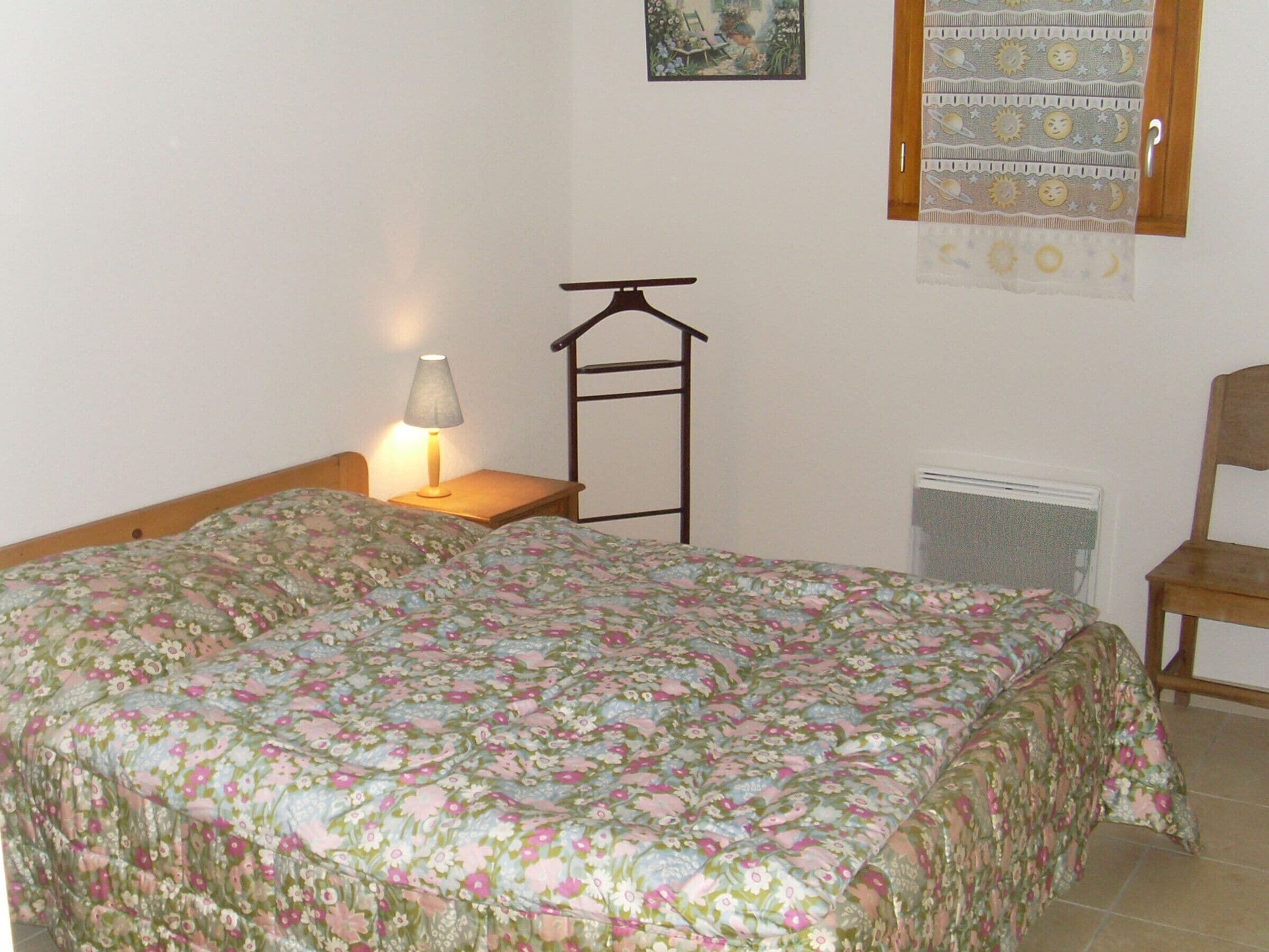 A bedroom with a double bed, a bedside table with a lamp, a wooden chair, a radiator, and a window with a curtain.
