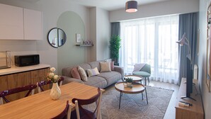 A modern living room with a sofa, dining table, chairs, a TV, and a large window with curtains. - Cozy Retreat: Stylish 1BR Apt (Esenyurt)