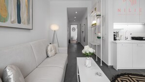 A modern, minimalist living room with a white sofa, a coffee table, and a painting on the wall. A hallway leads to a kitchen area with white cabinets and a sink. - Lusso Bianco Nilie Hospitality MGMT (Thessaloniki)