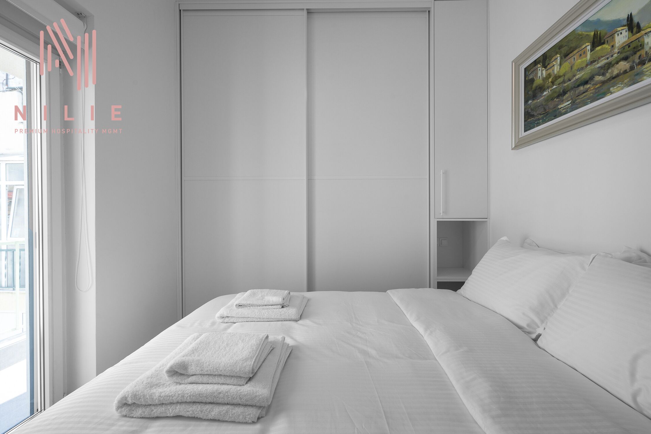Une chambre d’hôtel avec un lit, du linge de lit blanc, un tableau au mur et une fenêtre donnant sur un beau paysage.
