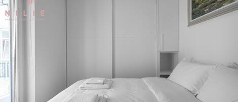 Une chambre d’hôtel avec un lit, du linge de lit blanc, un tableau au mur et une fenêtre donnant sur un beau paysage.