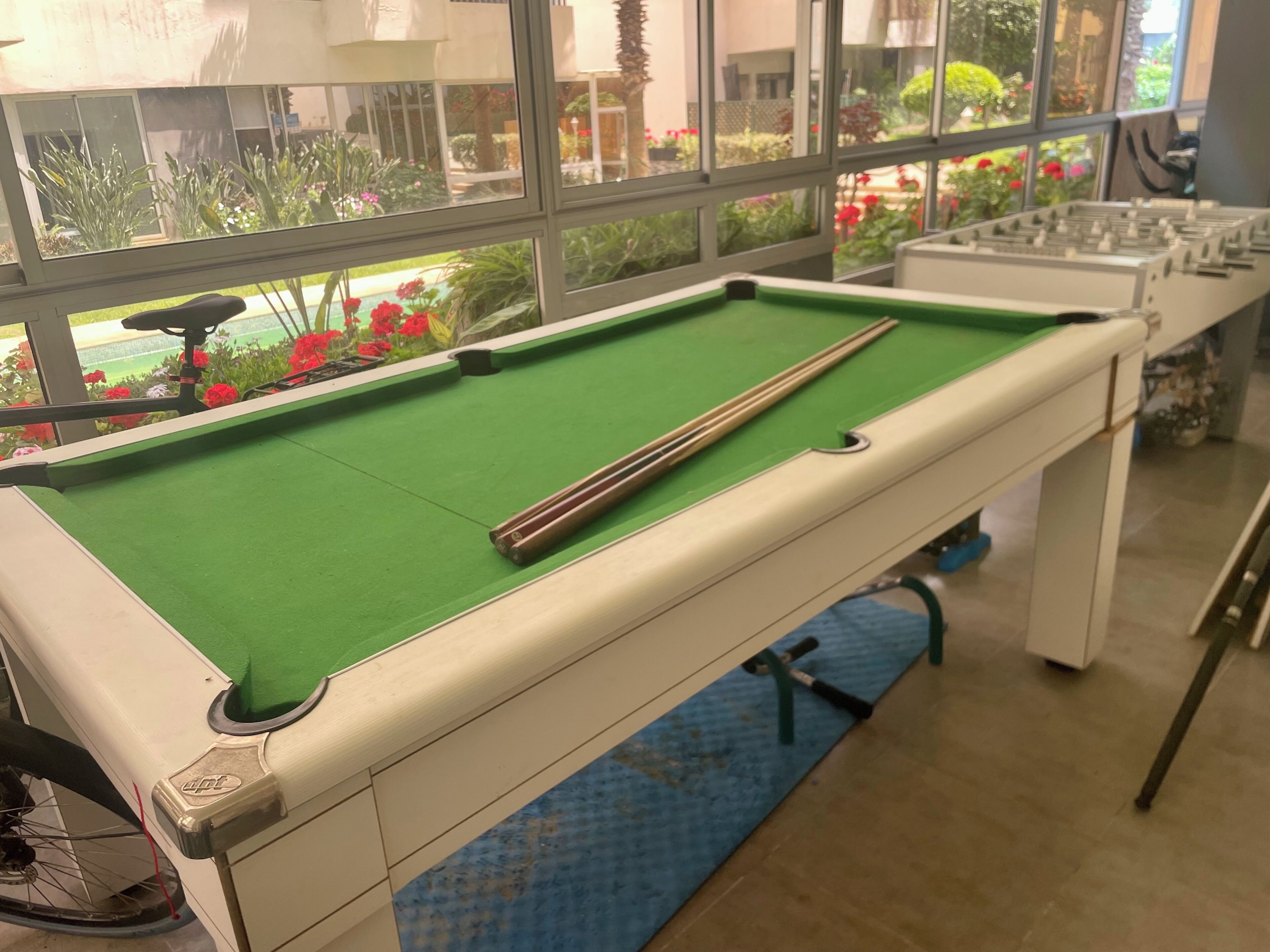 Una mesa de billar con taco largo, una ventana que da a un jardín y una mesa de futbolín al fondo.