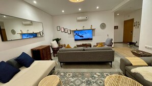 Un salon moderne avec un canapé gris, une table basse en bois et un téléviseur fixé au mur. On y trouve un grand miroir au-dessus du canapé, une plante en pot et un fauteuil en osier.