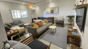 Un salon moderne avec un canapé gris, une petite table et un fauteuil. Une fenêtre offre une vue sur la verdure à l’extérieur.