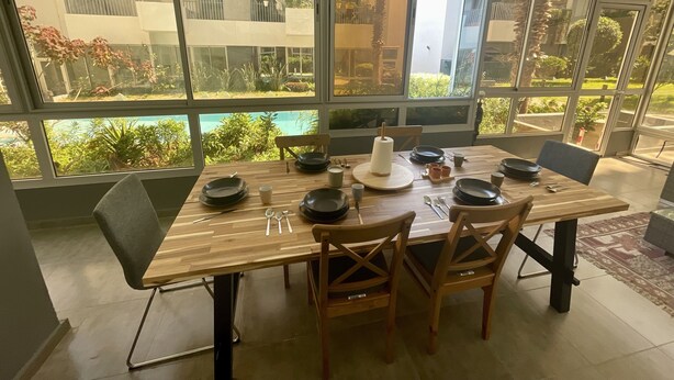 Un coin repas avec une table en bois dressée pour quatre personnes, entouré de grandes fenêtres donnant sur un immeuble résidentiel et sur la verdure.