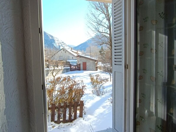 A view through a door of a snowy landscape with a wooden fence, a building, and mountains in the background. - Cozy 1BR Apartment with Private Garden, 300m to Pistes in Briançon (Briançon)