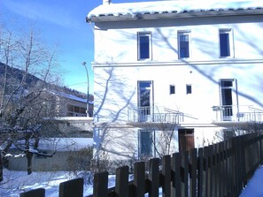 Un edificio de varios pisos con un balcón, una valla de madera en primer plano y suelo cubierto de nieve.