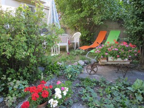 Un jardín con plantas en macetas, un carretón y mobiliario exterior.