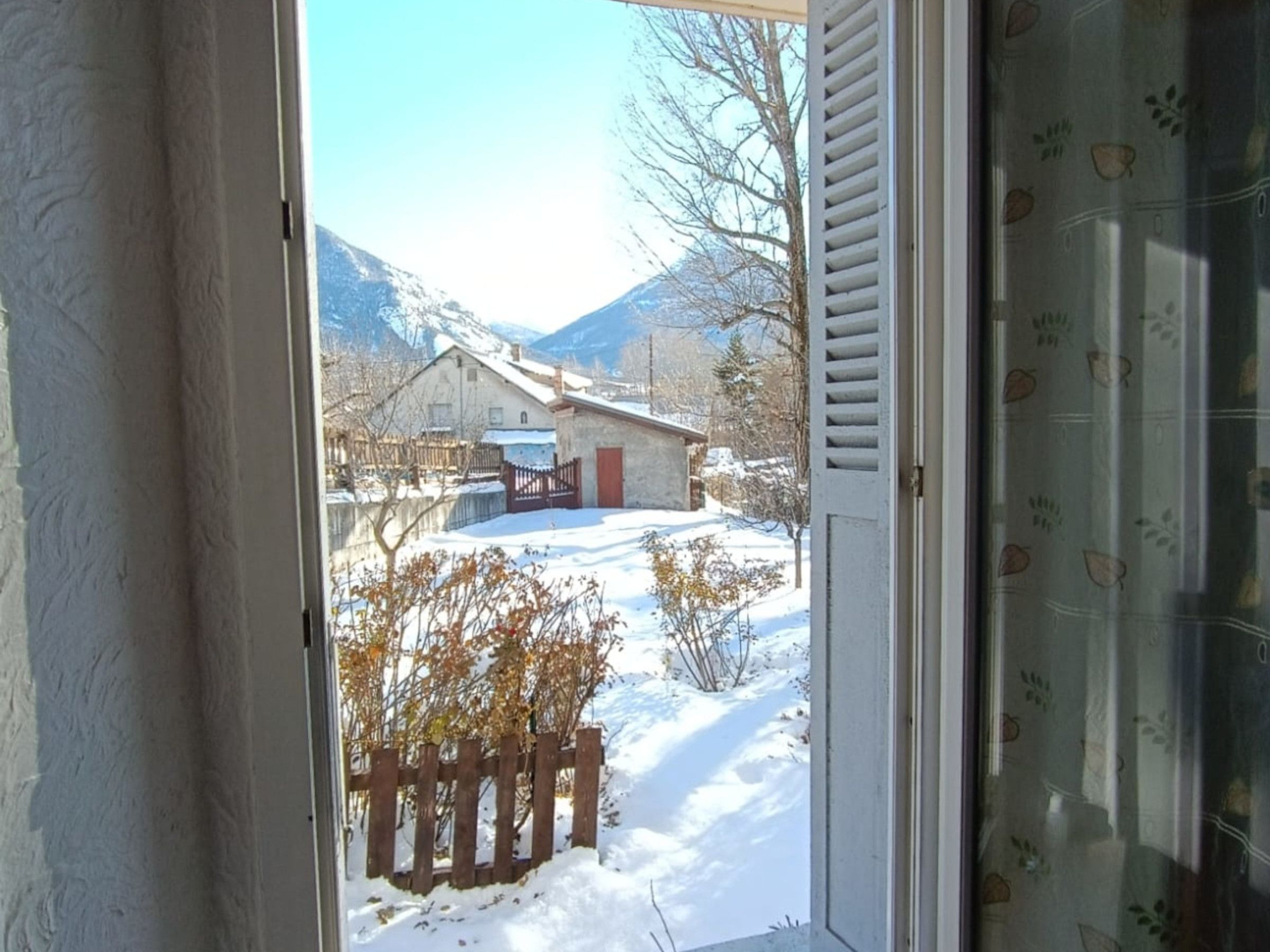 Une vue à travers une porte, donnant sur un paysage enneigé avec une clôture en bois, un bâtiment et des montagnes en arrière-plan.