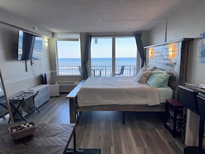 Quarto com cama, televisão, escrivaninha, cadeira e vista para o mar.