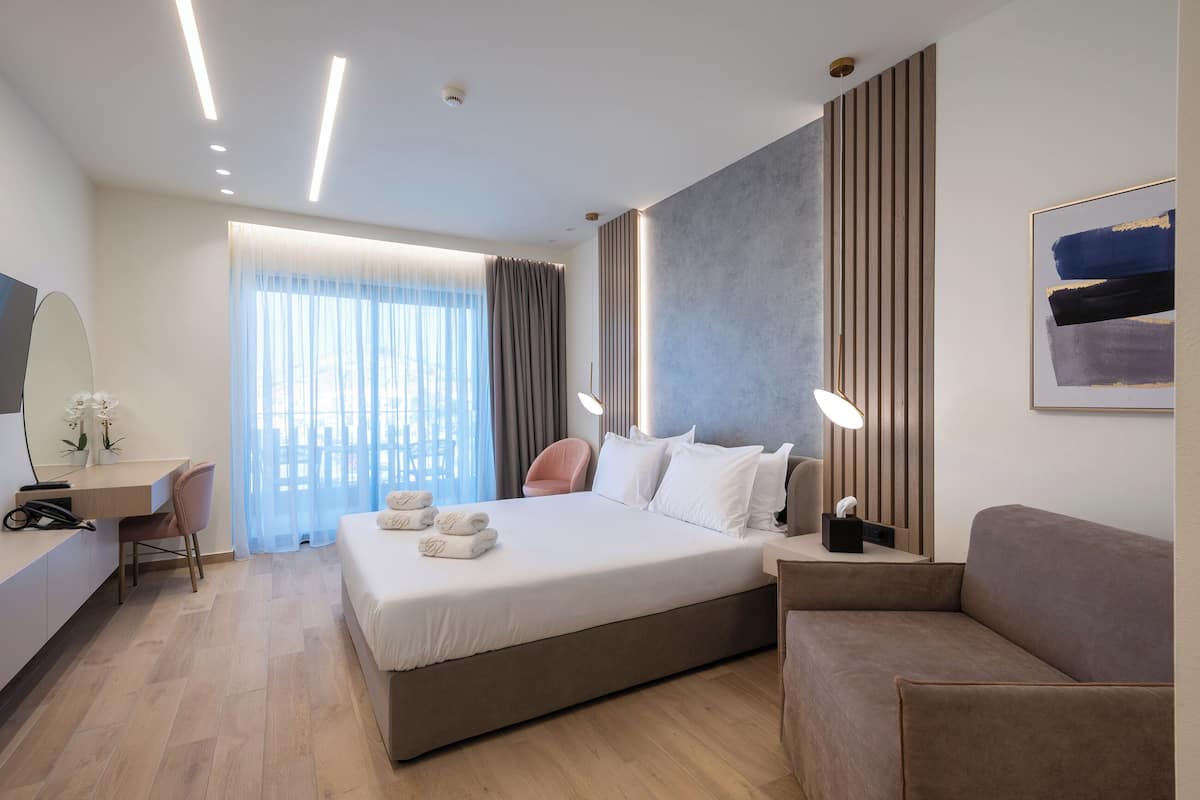 A modern hotel room with a large bed, a desk, a chair, a sofa, and a large window with curtains.