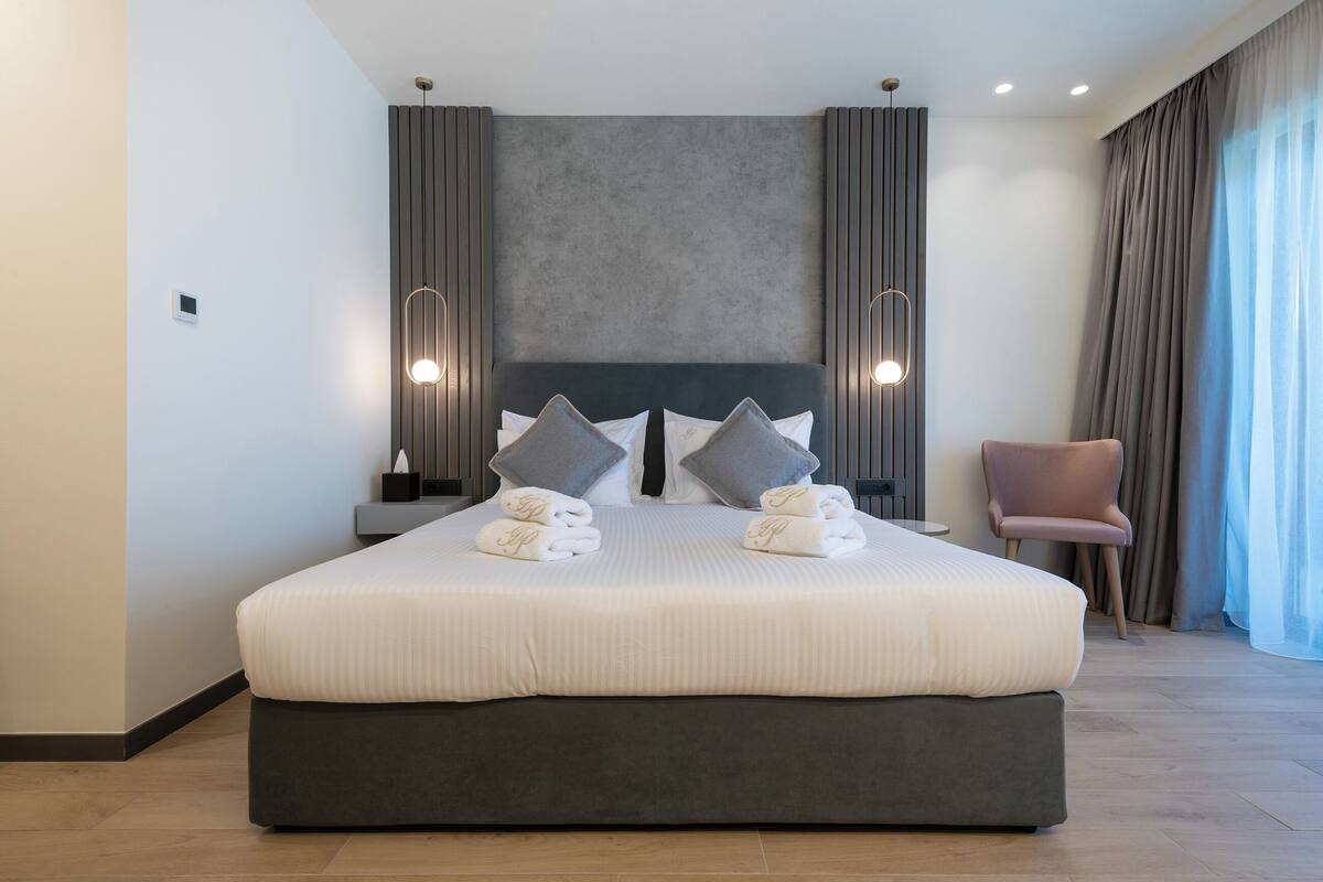 A modern hotel room with a large bed, two bedside tables, a chair, and a view of the outdoors.