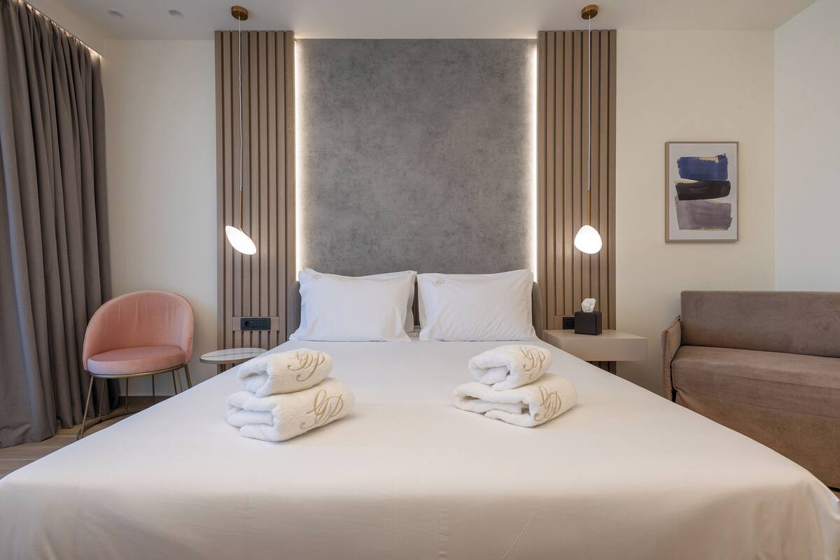 A hotel room with a bed, two pillows, a nightstand, a chair, a sofa, and towels on the bed.