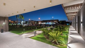 A modern building with a flat roof, a paved courtyard with palm trees, and a view of a cityscape at dusk. - Grand Patelli (Lemnos)