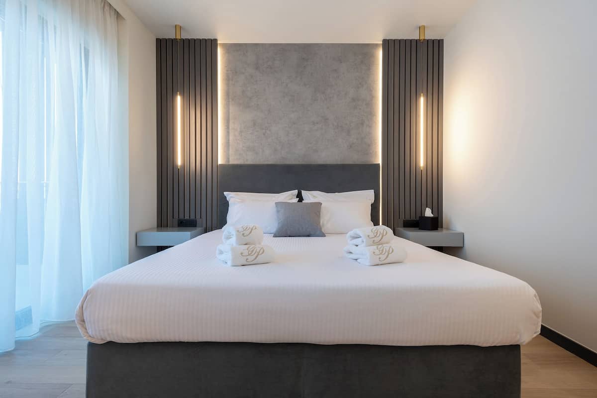 A modern hotel room with a large bed, bedside tables, a headboard, and a view of the outdoors.