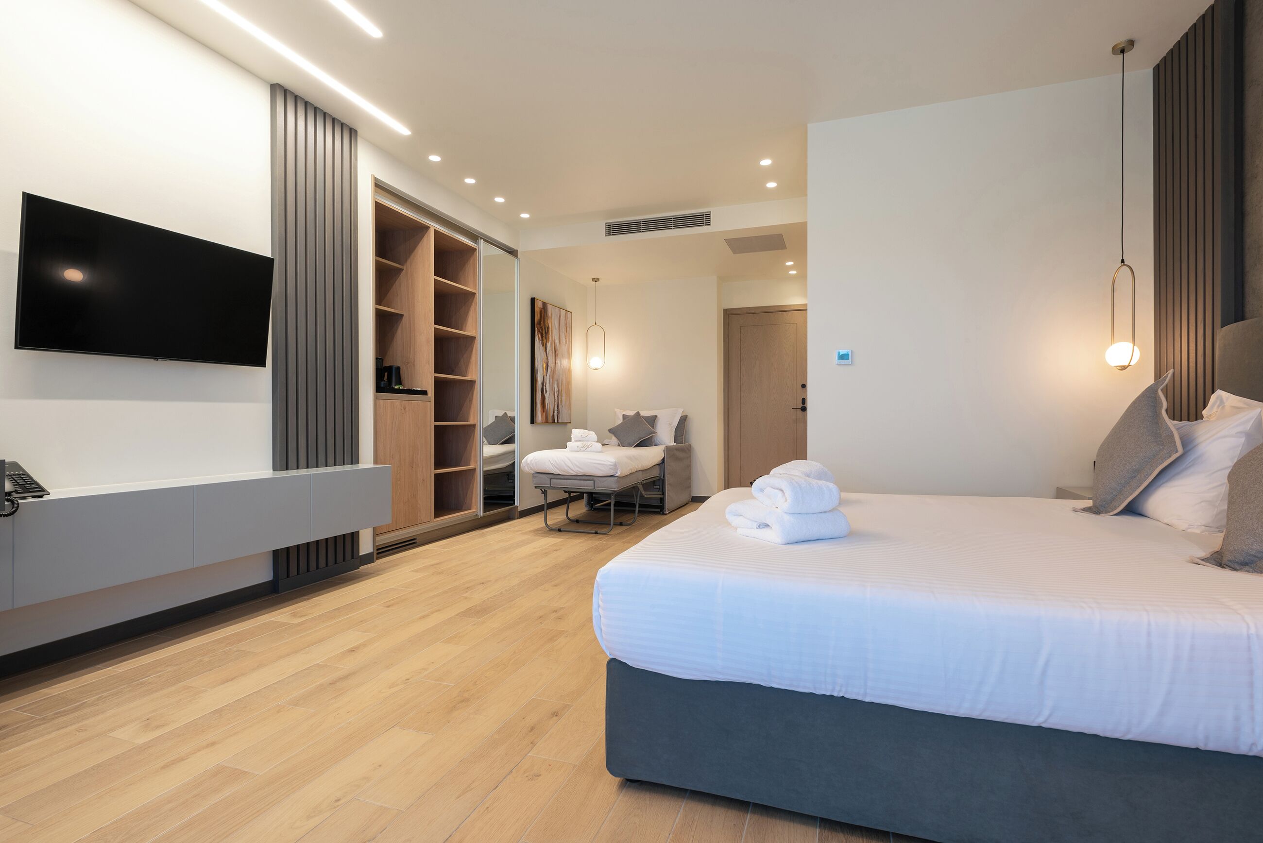 Une chambre d’hôtel moderne dotée d’un grand lit, d’un téléviseur à écran plat fixé au mur, d’un dressing et d’une salle de bain avec douche et toilettes.