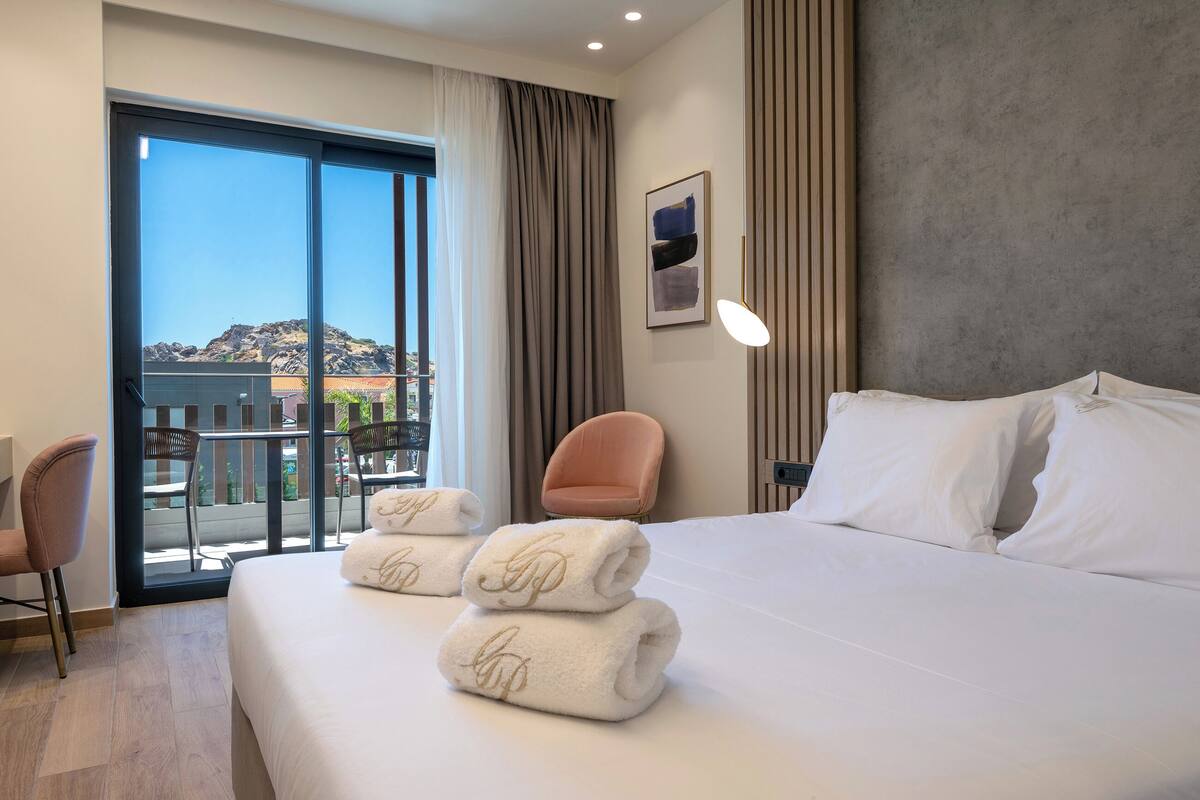 A hotel room with a bed, a chair, a desk, and a view of a cityscape through large windows.