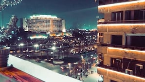 A cup of coffee on a wooden table with a view of a cityscape at night, including a building with illuminated windows and a busy street below. - Rotana Palace Suites (Cairo)