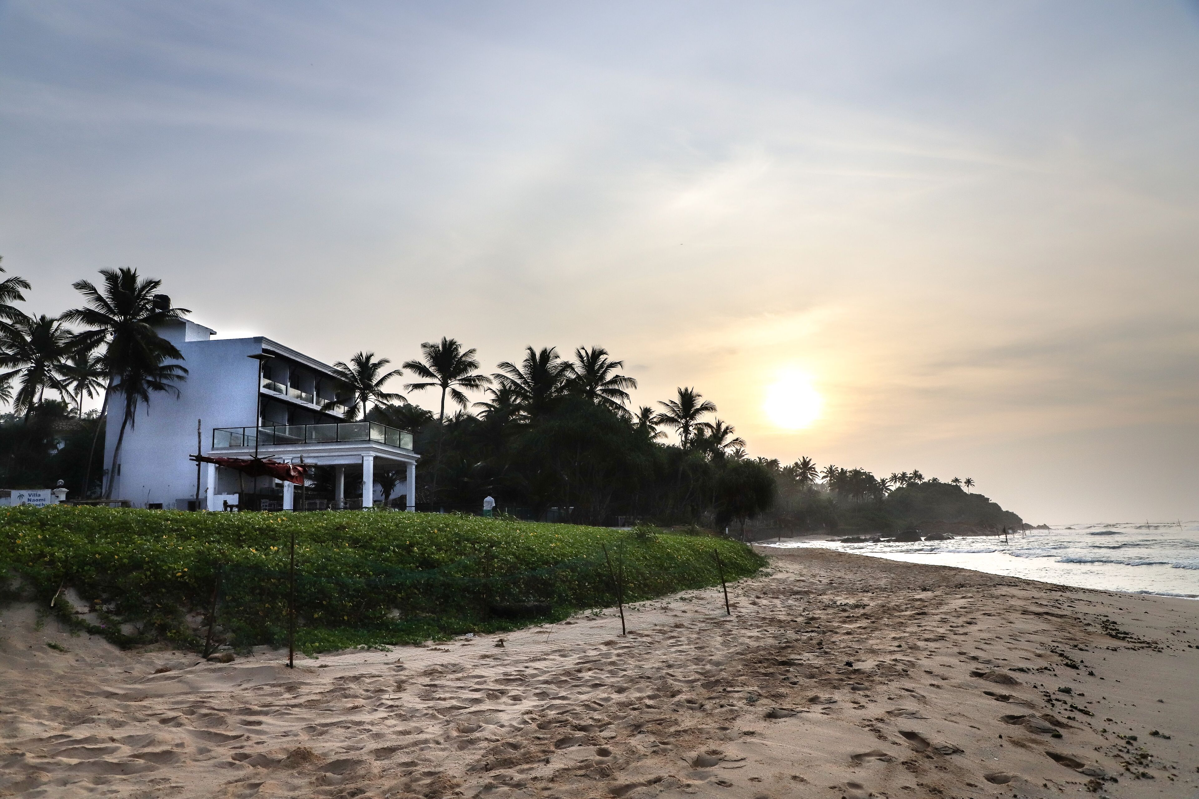 Una propiedad frente a la playa con un edificio blanco, palmeras y una playa de arena.